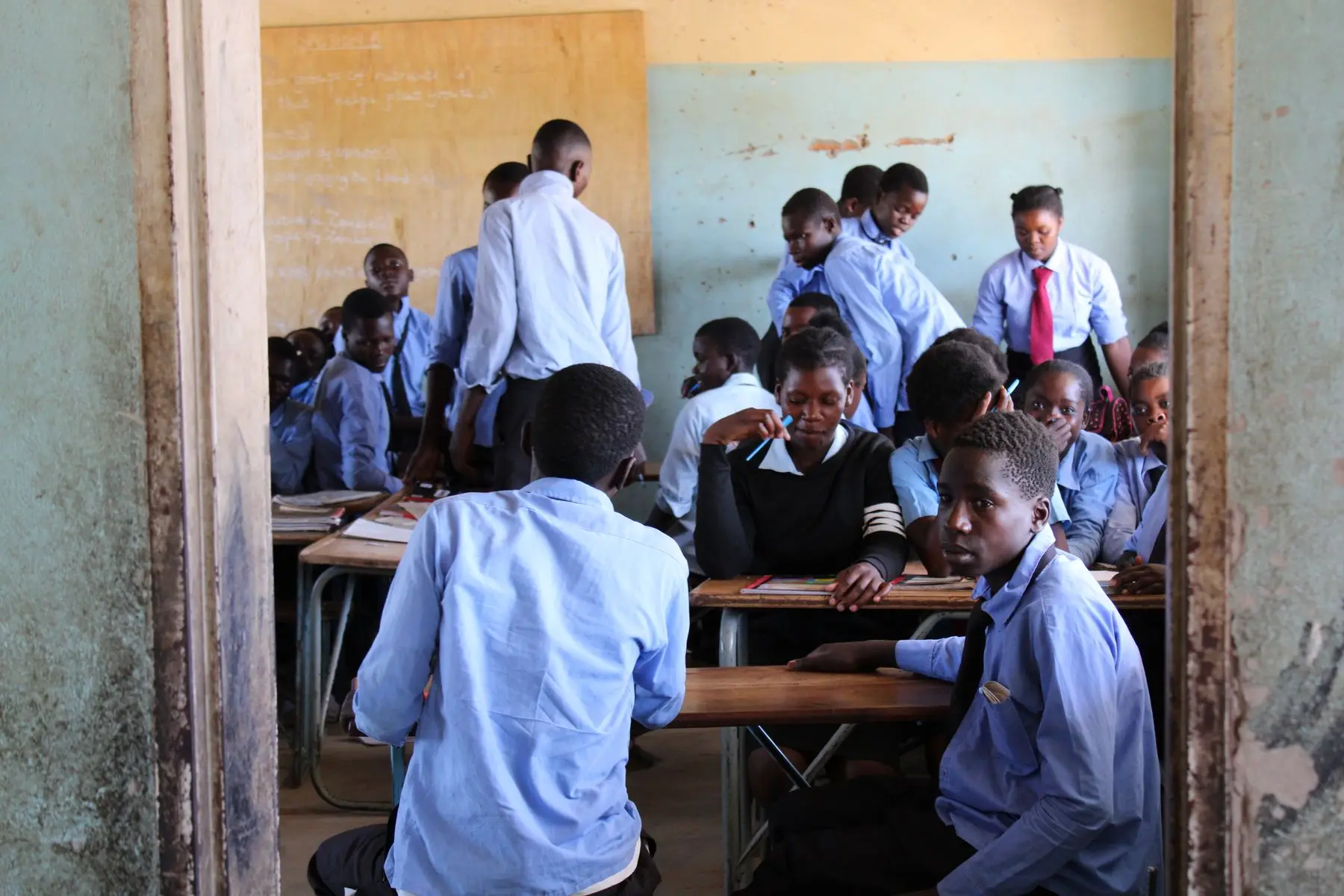 A busy secondary school classroom in East Africa seen from the doorway, learners at shared wooden desks